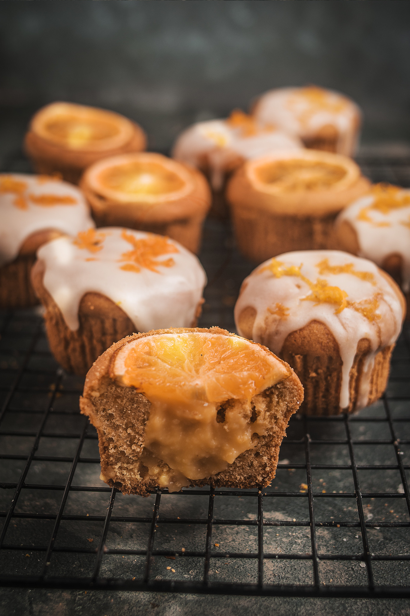 Cut-open blood orange cupcake revealing a soft curd-filled centre, topped with a baked slice of orange, with iced cupcakes blurred in the background.